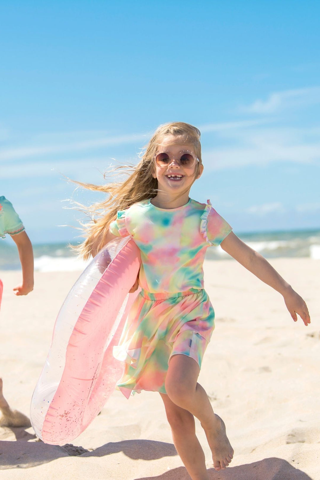 A young girl with blonde hair and sunglasses runs on a sandy beach carrying a pink inflatable pool float. She is wearing a pastel tie-dye dress with pink, green, and blue colors, with the blue ocean and sky in the background.