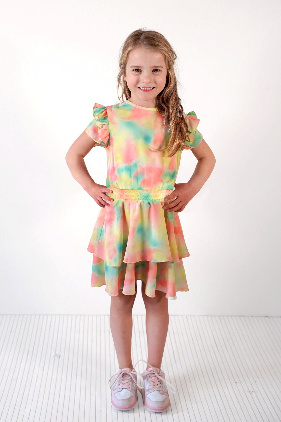 A young girl with blonde hair stands with hands on hips wearing a pastel tie-dye ruffled dress and light pink sneakers against a white studio background.