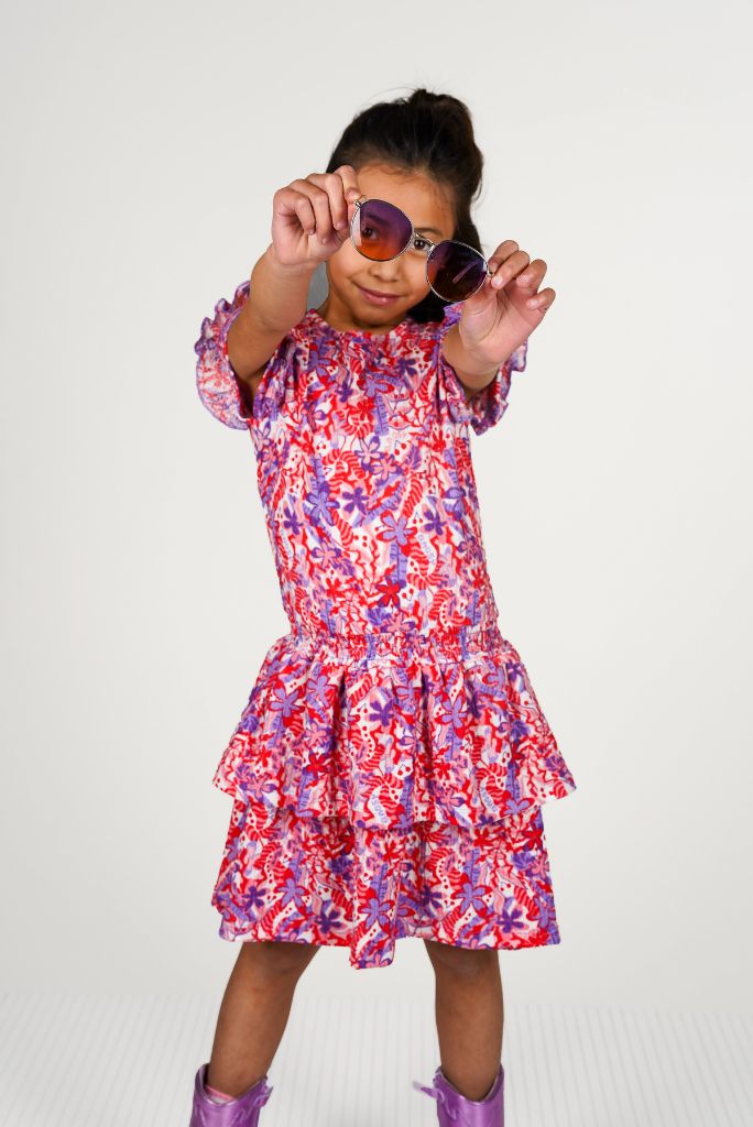 Young girl wearing the colorful floral dress Delphi holding sunglasses against a white background