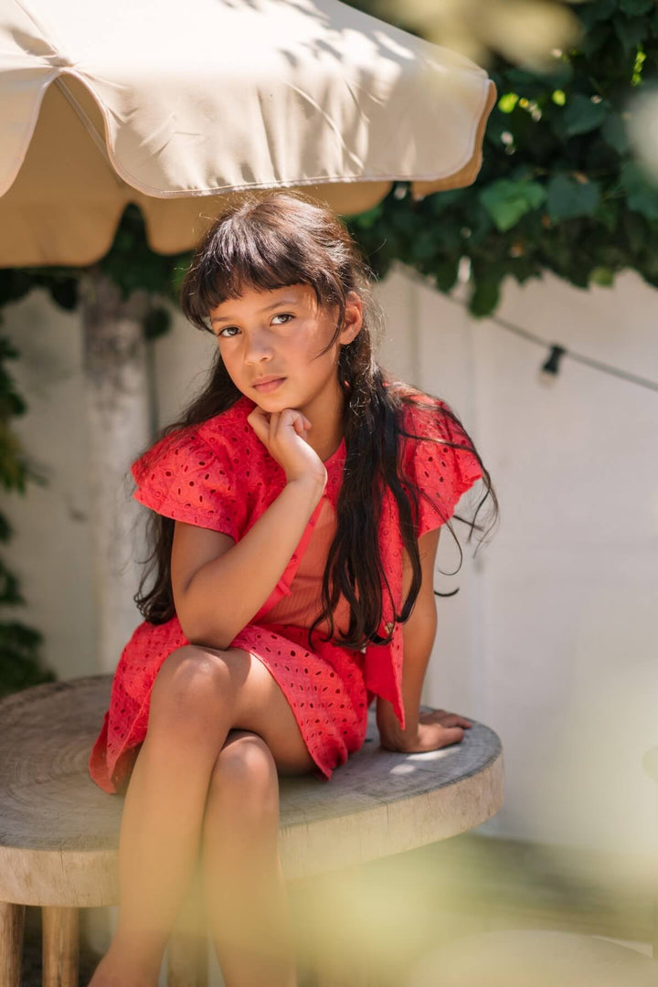 Young girl in a coral pink blouse and skirt sitting under an umbrella with greenery in the background