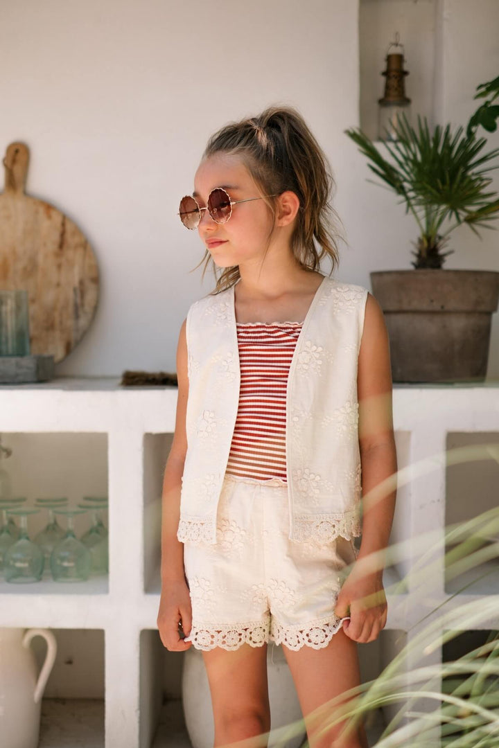 Young girl wearing a striped singlet and white vest with shorts standing in a casual setting.