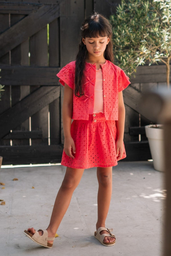 A young girl wearing a bright coral pink two-piece outfit consisting of a short-sleeve button-front top and matching mini skirt, paired with beige sandals, standing outdoors in front of a dark wooden fence.