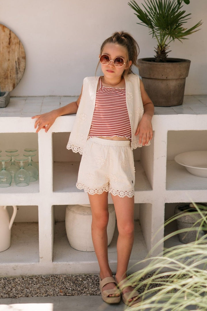 A young girl wearing a matching cream lace-trimmed vest and shorts set with a red-and-white striped top, standing in front of a white tiled outdoor counter.