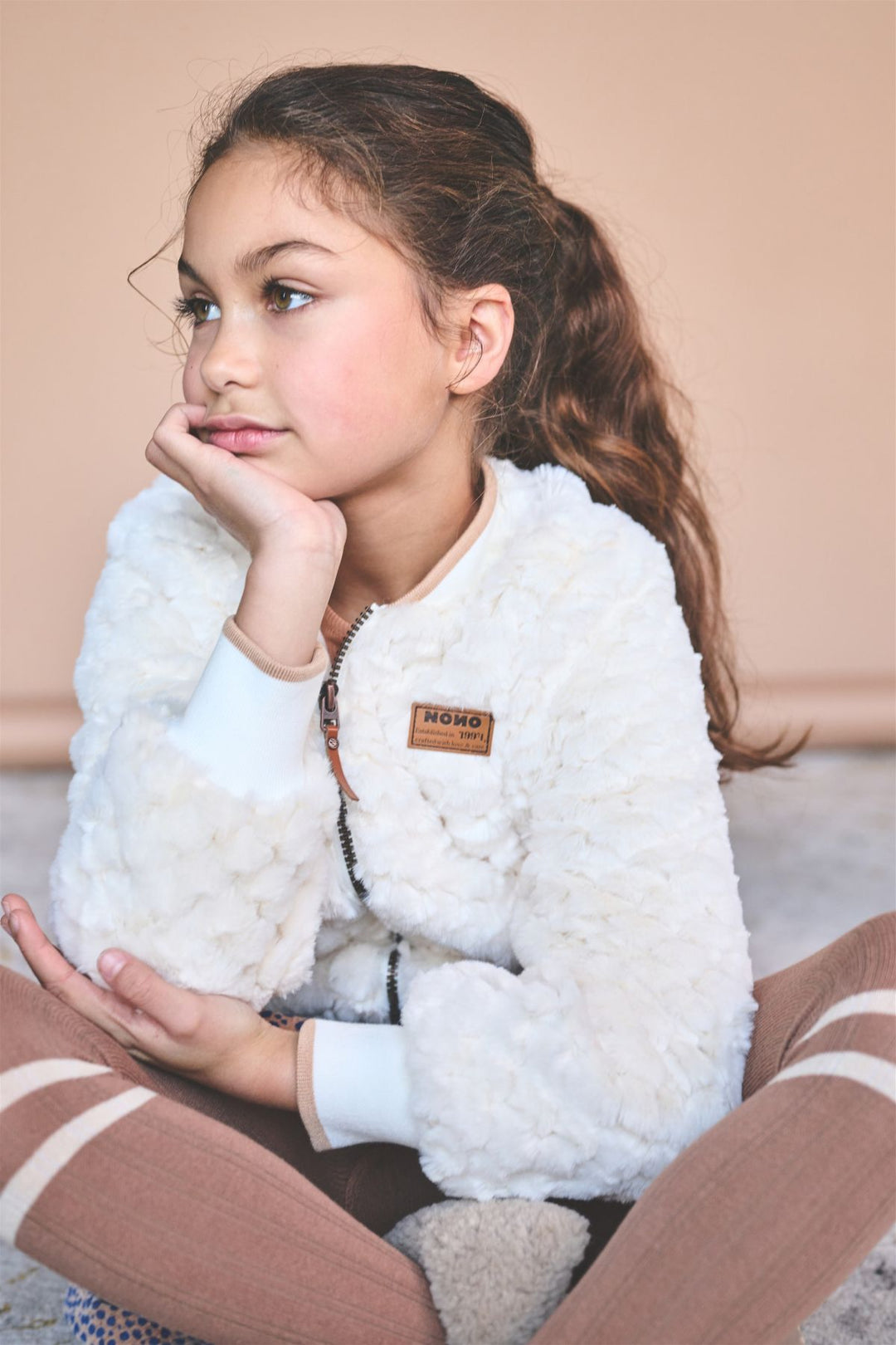 A young girl with brown hair in a ponytail sits cross-legged, wearing a white fuzzy zip-up jacket with a brown &