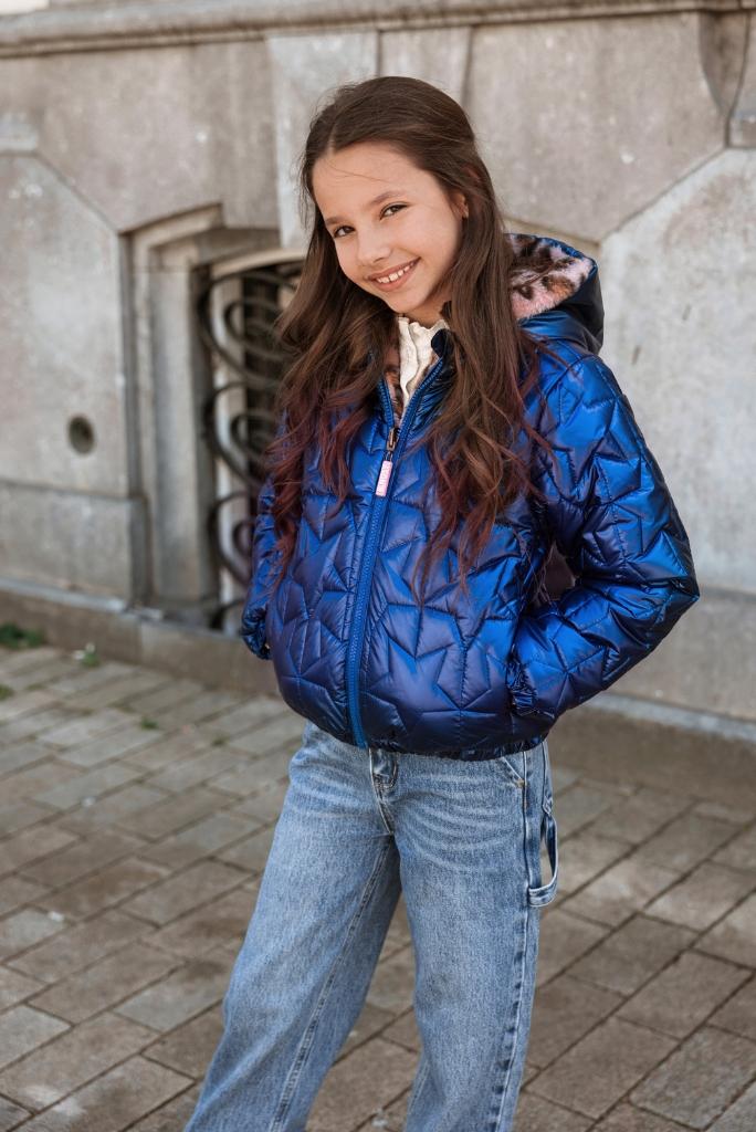 Young girl wearing a blue puffer jacket standing on a stone pavement.