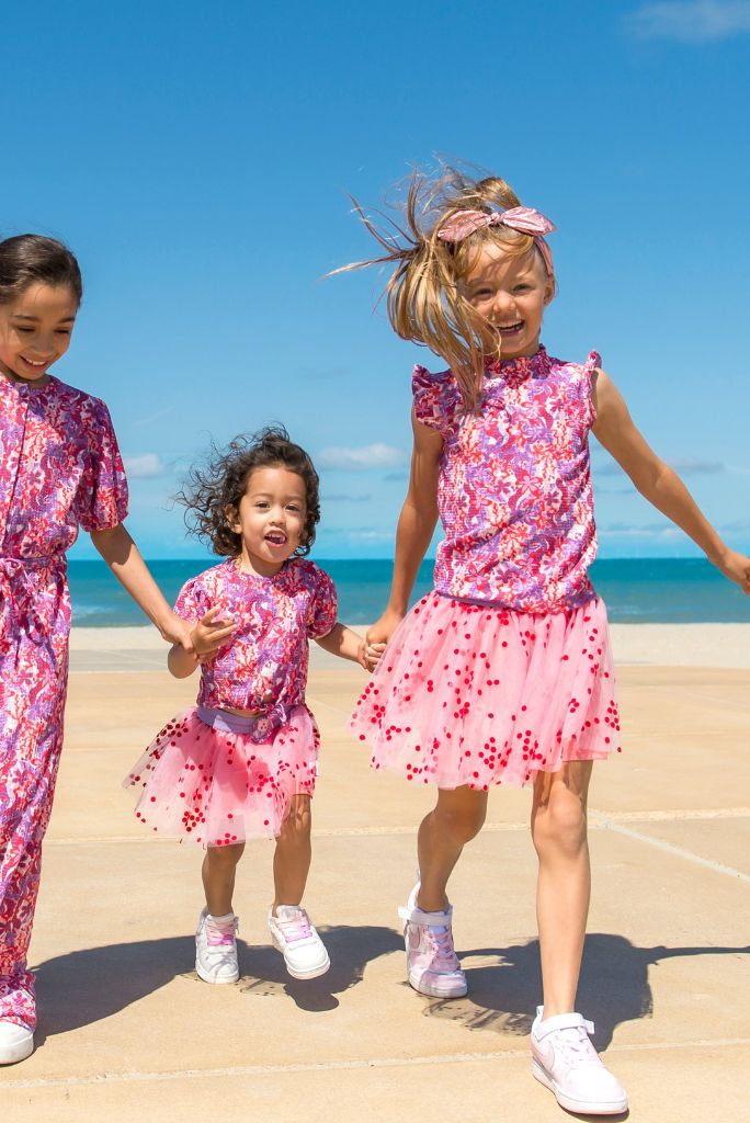 Three children in matching pink outfits standing on a beach with blue sky and ocean in the background.