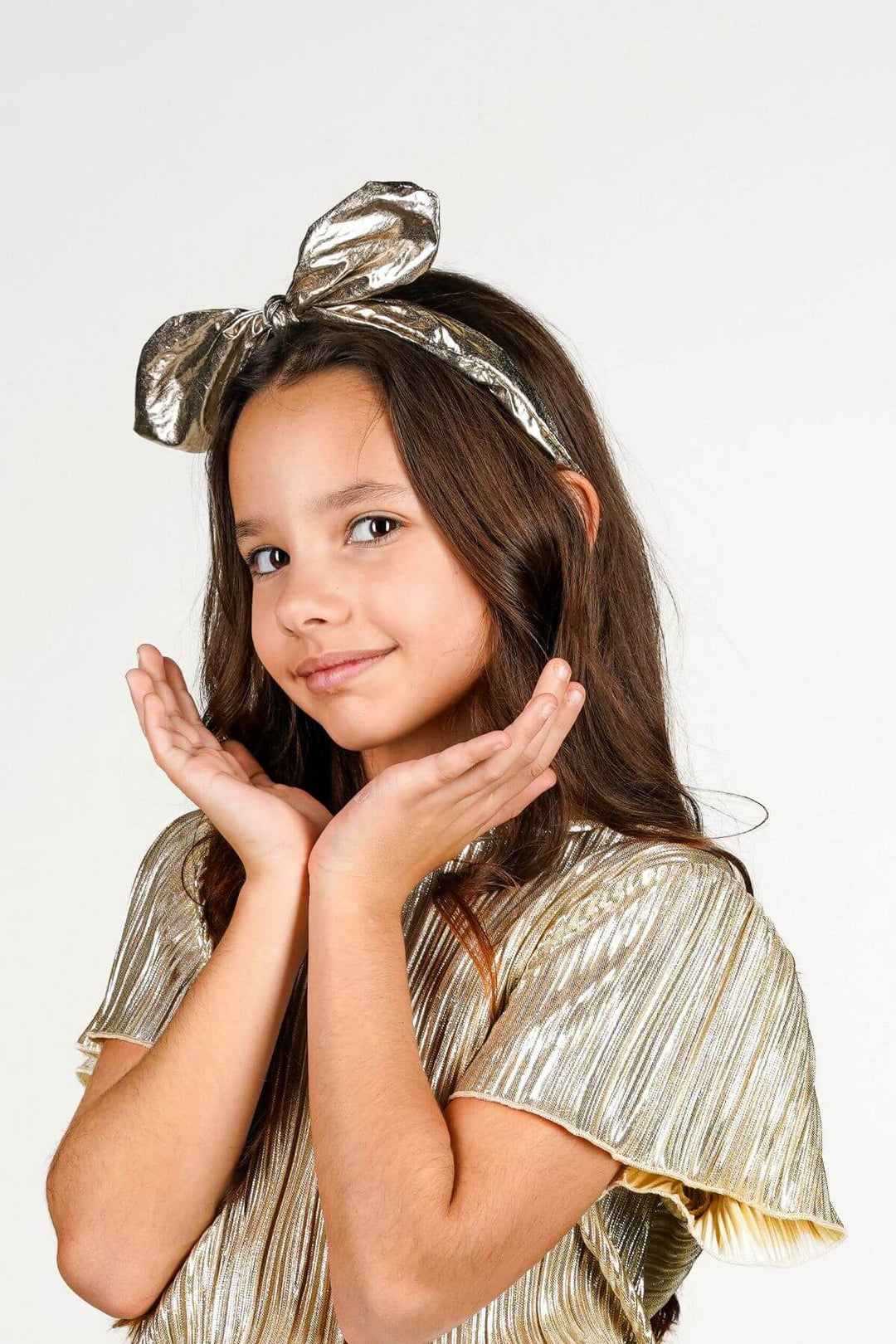 A young girl with long brown hair wearing a shiny metallic gold bow headband and a matching pleated gold top, posing with her hands near her face against a plain white background.