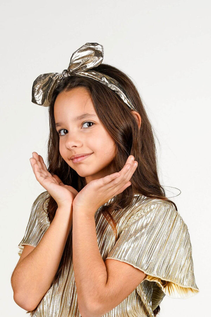 A young girl with long brown hair wearing a shiny metallic gold bow headband and a matching pleated gold top, posing with her hands near her face against a plain white background.