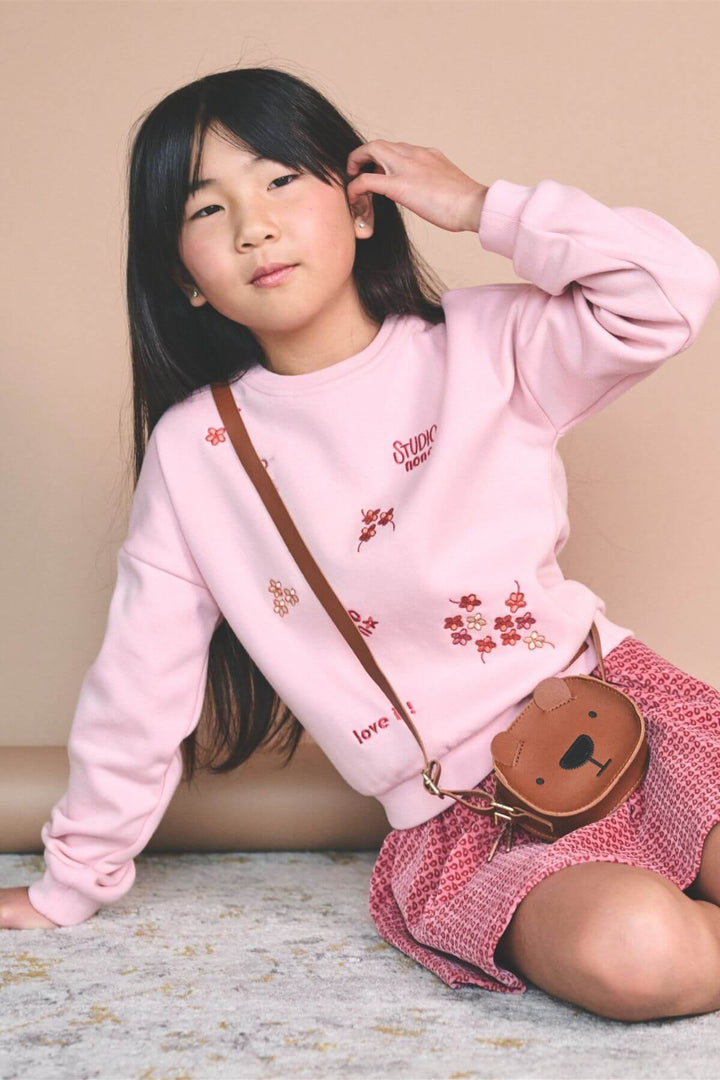 A young girl with long black hair sits on a patterned rug wearing a light pink sweatshirt with red floral embroidery and a pink-and-white checkered skirt. She is carrying a brown teddy bear-shaped crossbody bag with a brown strap.