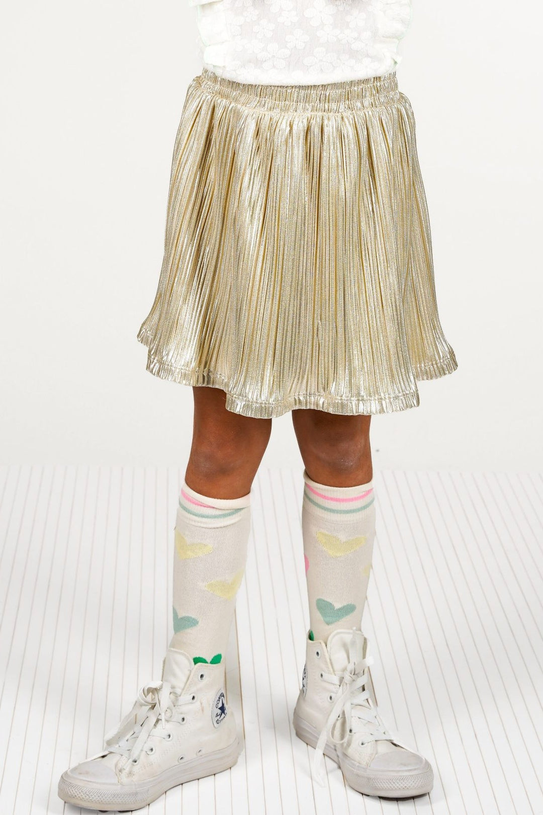 A child wearing a shiny metallic gold pleated skirt, white knee-high socks with pastel heart patterns, and white high-top sneakers.