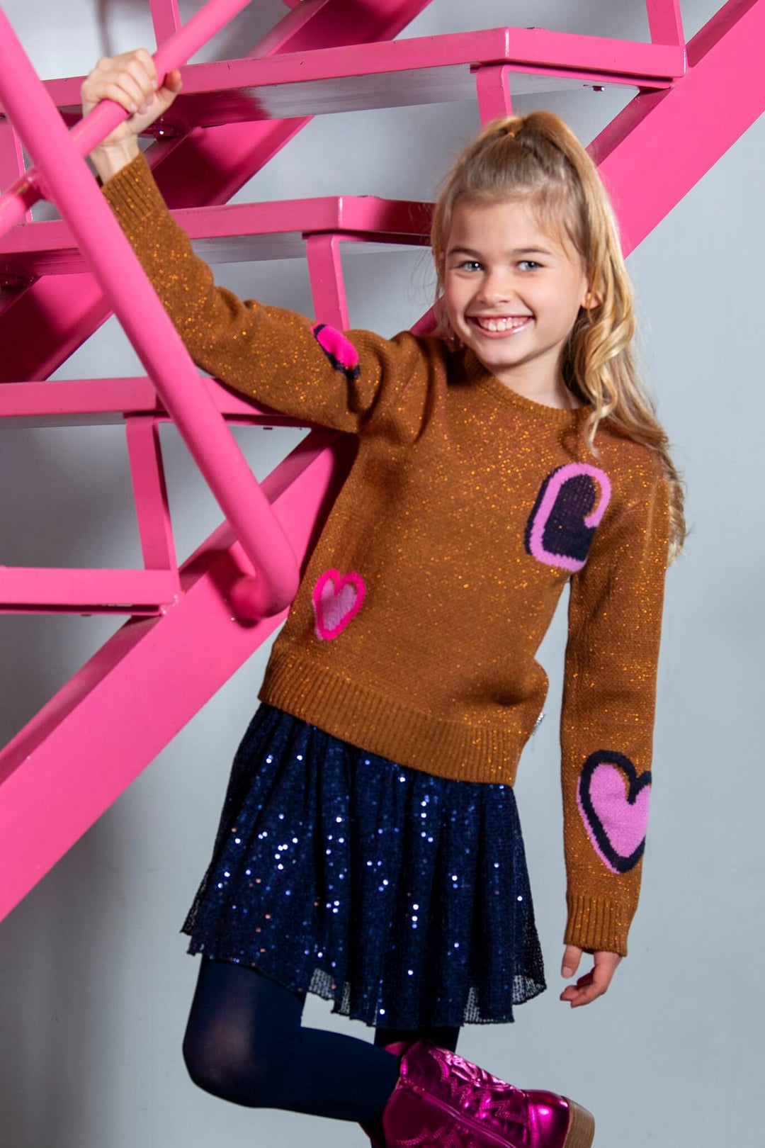 Young girl wearing a brown jumper with pink heart designs and a blue skirt, standing against a pink metal structure.