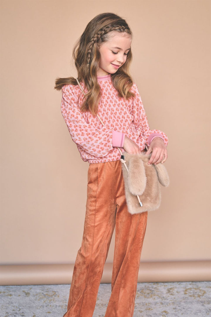A young girl with long brown hair in braids is wearing a pink patterned long-sleeve top and rust-orange wide-leg pants, holding a beige faux-fur clutch bag against a solid beige studio backdrop.