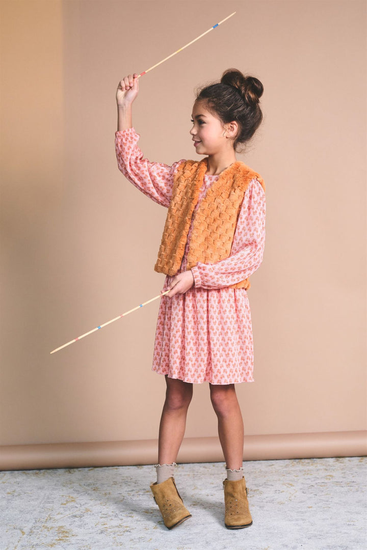 A young girl with her hair in a bun stands in a studio wearing a pink patterned long-sleeve dress, an orange textured vest, and tan ankle boots, holding a long wooden pointer wand.