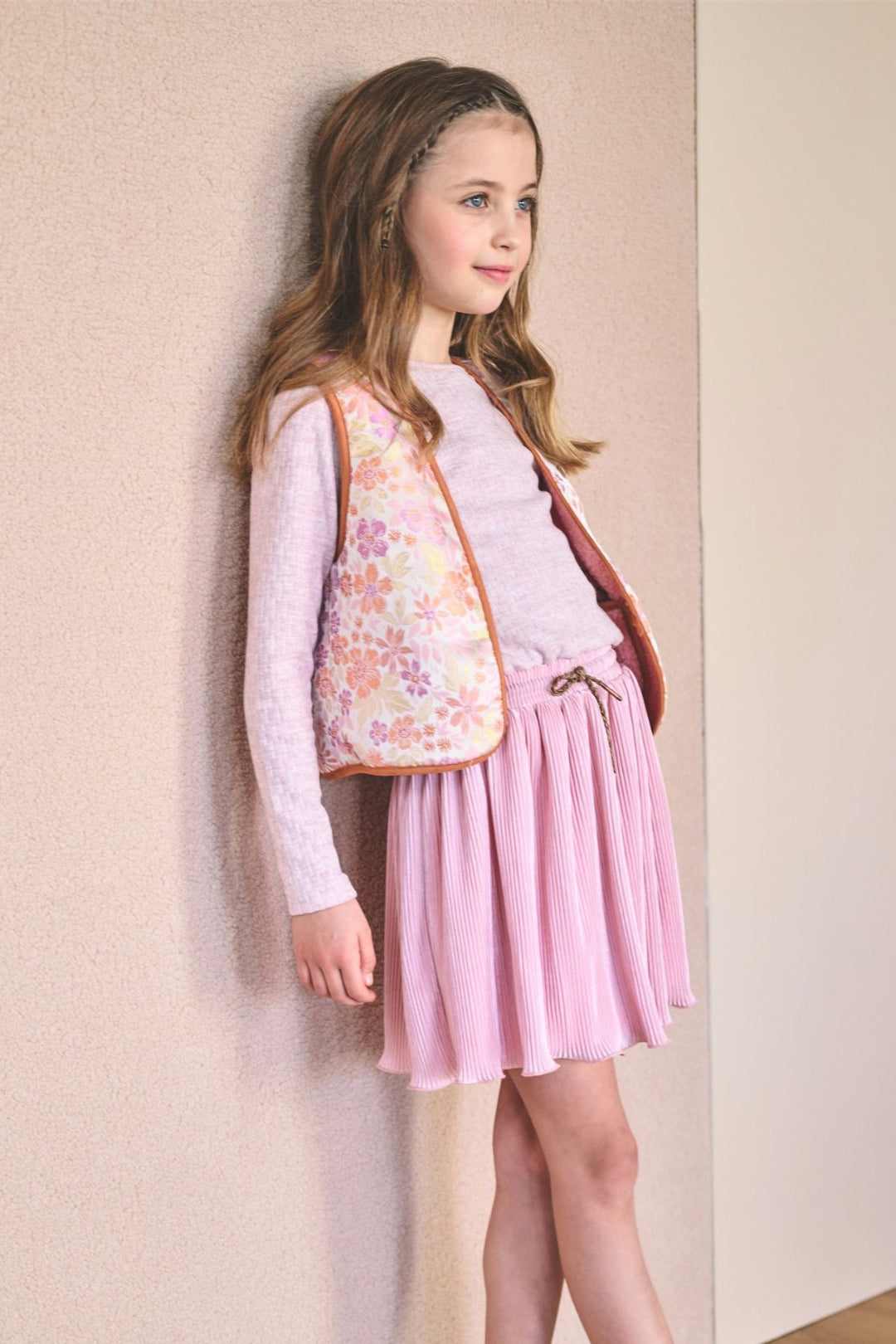 A young girl stands against a textured beige wall wearing a light pink long-sleeve top, a pleated pink skirt, a floral-patterned vest, white socks, and brown suede shoes.