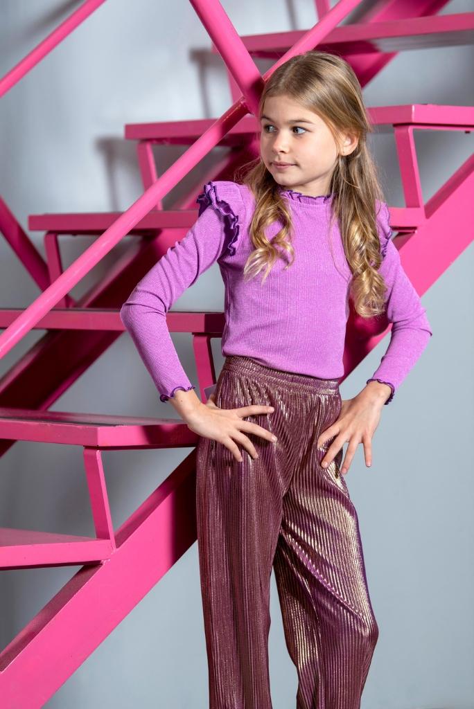 Young girl in a pink top and maroon pants standing against a pink geometric background
