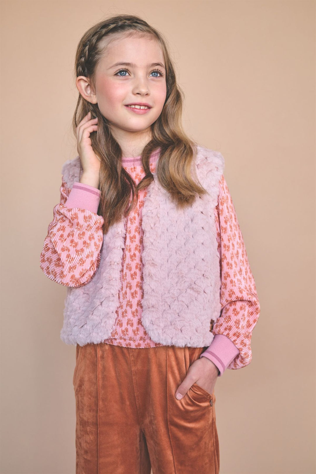 A young girl with long light brown hair wearing a pink patterned long-sleeve blouse, a fuzzy light pink vest, and rust-brown pants, posing against a solid beige studio background.