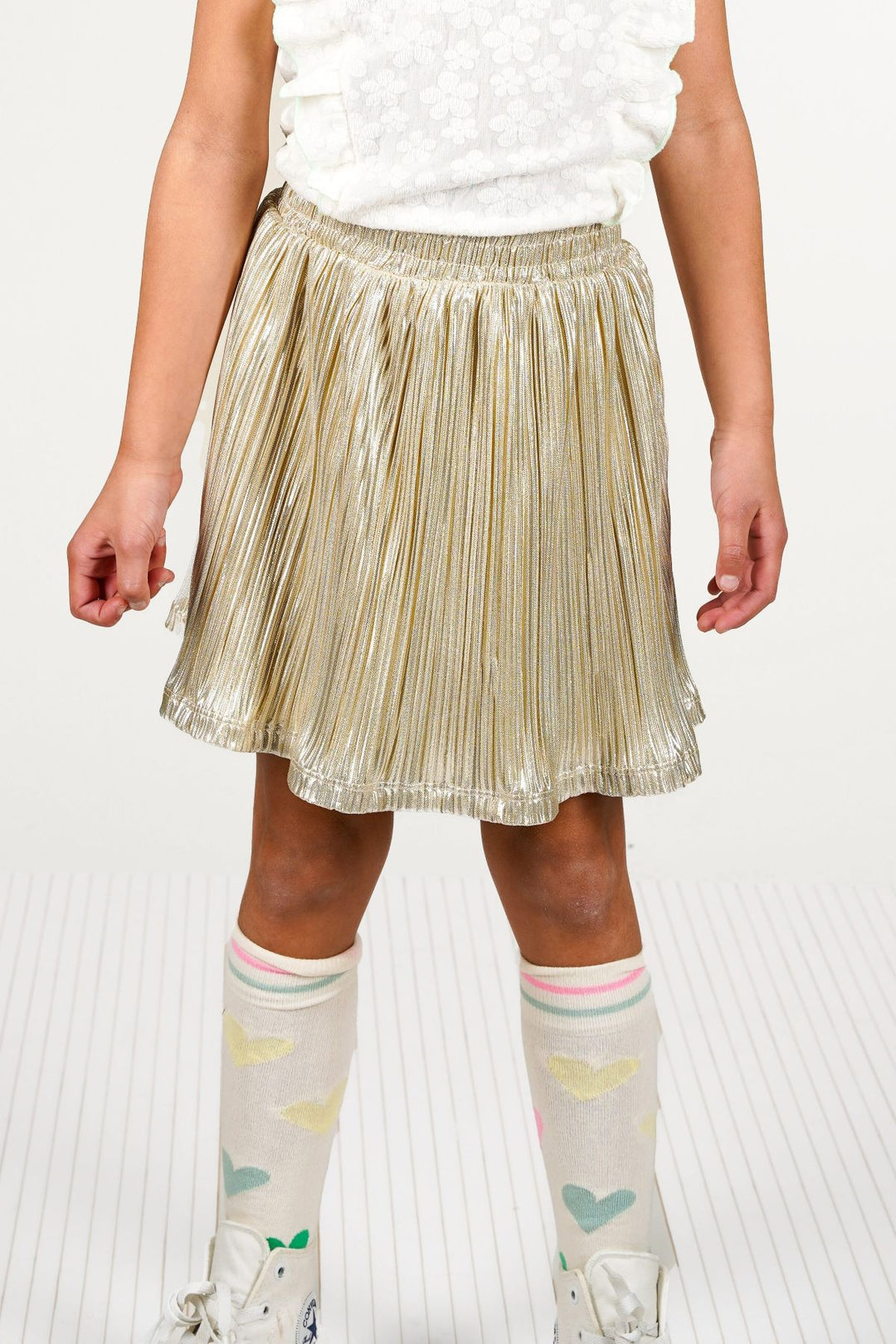 A girl wearing a shiny metallic gold pleated mini skirt, a white sleeveless top, and patterned knee-high socks with white sneakers, shown against a light studio background.