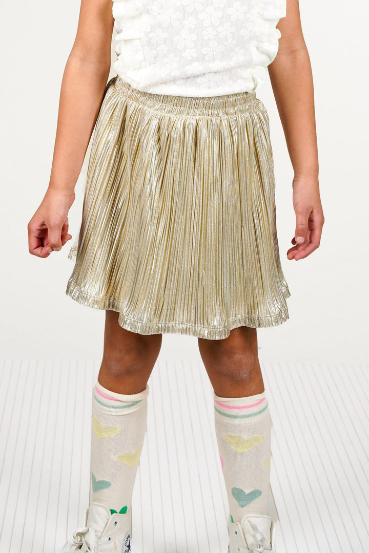 A girl wearing a shiny metallic gold pleated mini skirt, a white sleeveless top, and patterned knee-high socks with white sneakers, shown against a light studio background.