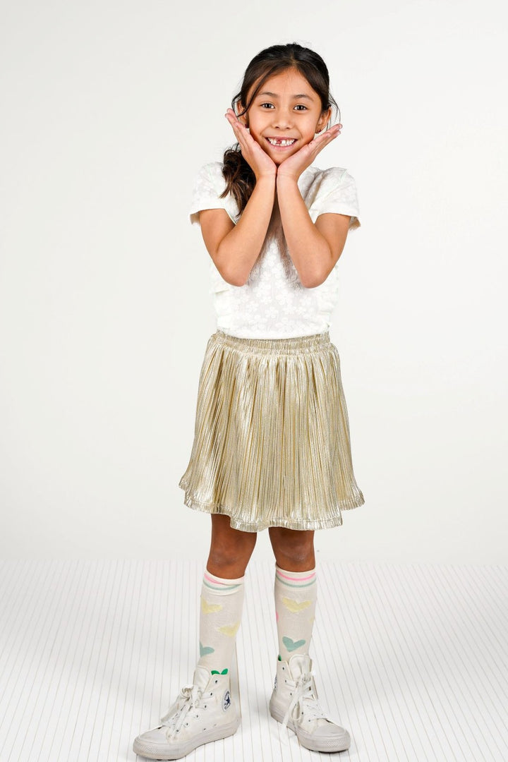 A young girl posing in a white short-sleeve top, a shiny metallic gold pleated skirt, patterned white knee-high socks, and white sneakers against a plain white studio background.