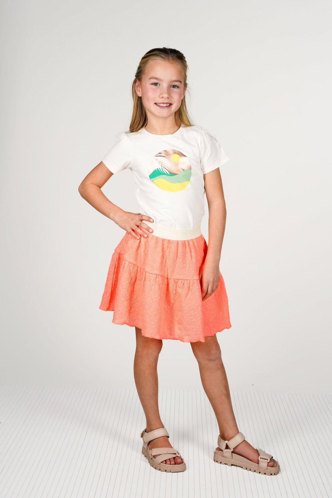 A young girl stands against a light gray studio background wearing a white short-sleeve graphic T-shirt with a colorful beach illustration and a tiered coral-orange skirt with beige platform sandals.