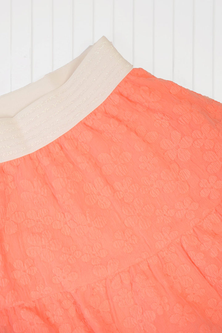 Close-up of a coral orange textured skirt with a cream elastic waistband, laid flat against a white paneled background.