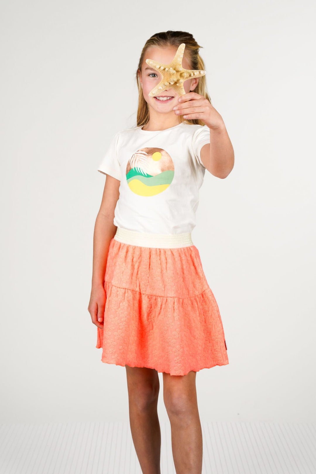 A young girl wearing a white graphic t-shirt and a tiered neon orange skirt. She holds a starfish in front of her eyes.