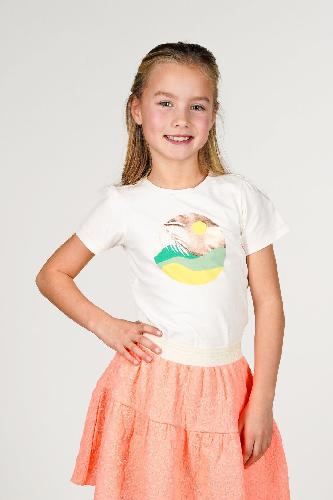 A young girl with blonde hair tied back, wearing a white short-sleeve t-shirt with a colorful graphic print and a tiered coral-pink skirt.