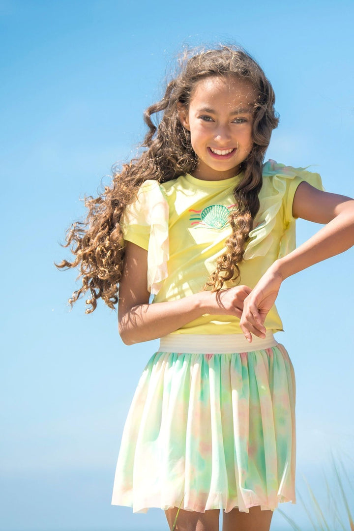 A young girl with long, curly brown hair smiles while wearing a yellow short-sleeve t-shirt and a pastel tie-dye pleated skirt against a bright blue sky background.