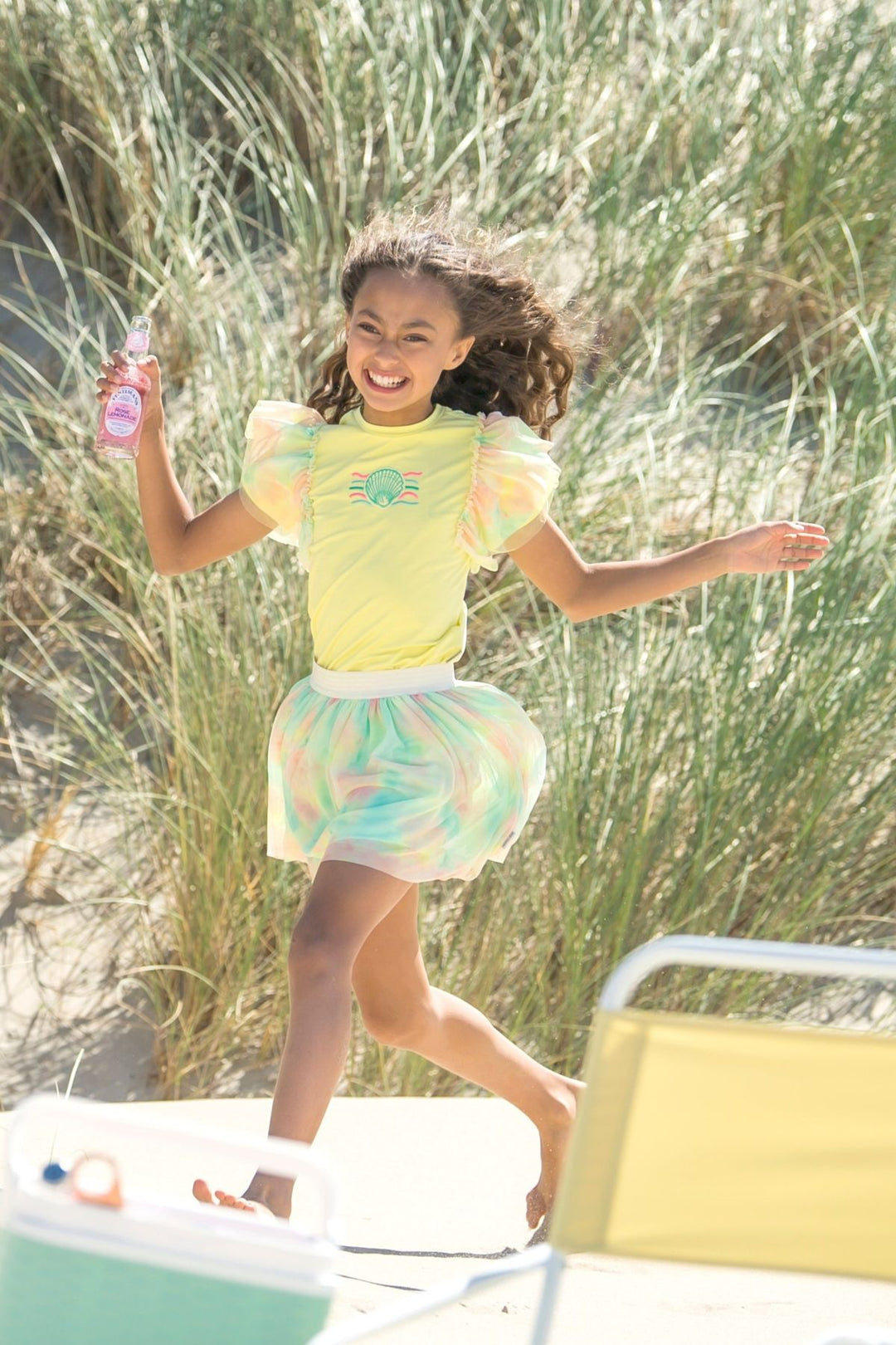 A young girl with curly brown hair runs and smiles in a beach dune setting, wearing a bright yellow short-sleeve top with puffy sleeves and a pastel multicolor tie-dye skirt. She is holding a pink bottle.