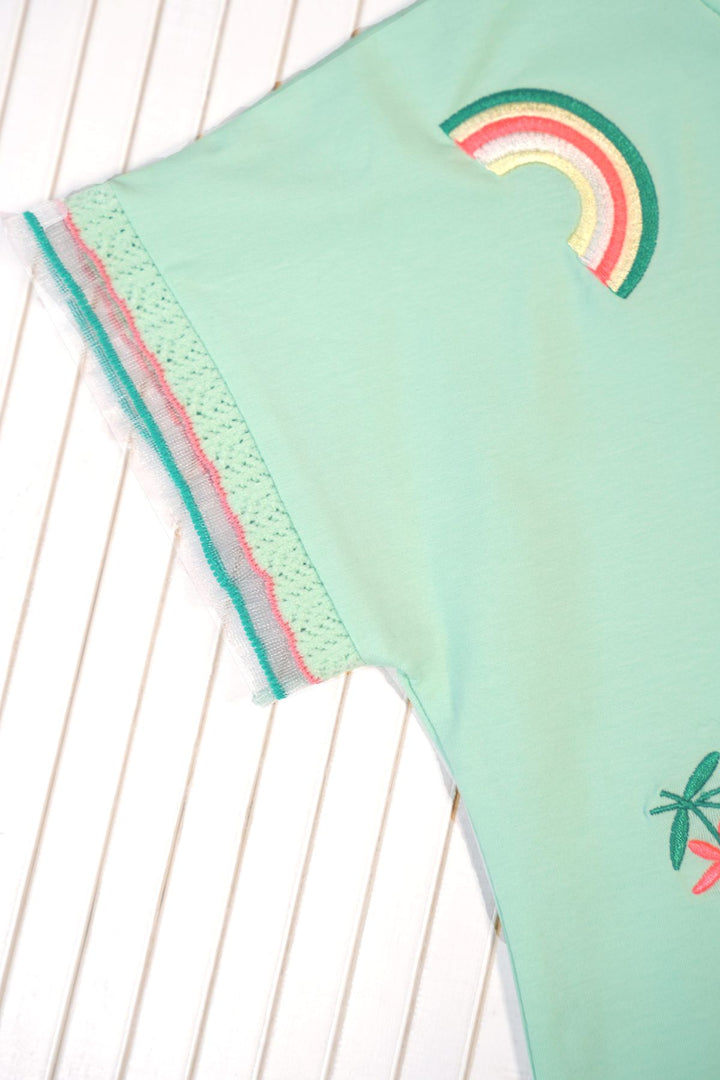 A close-up of a mint green garment laid flat on a white slatted surface, showing a rainbow patch, a palm tree motif, and a sheer lace trim with pink and teal edges.