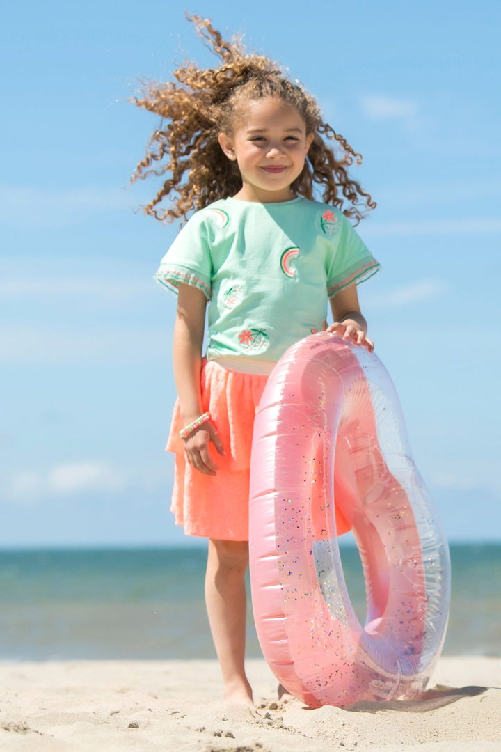 A young girl with curly hair stands on a sandy beach holding a pink and clear inflatable swim ring. She is wearing a mint green t-shirt with colorful tropical prints and a coral orange skirt, with the blue ocean and sky in the background.