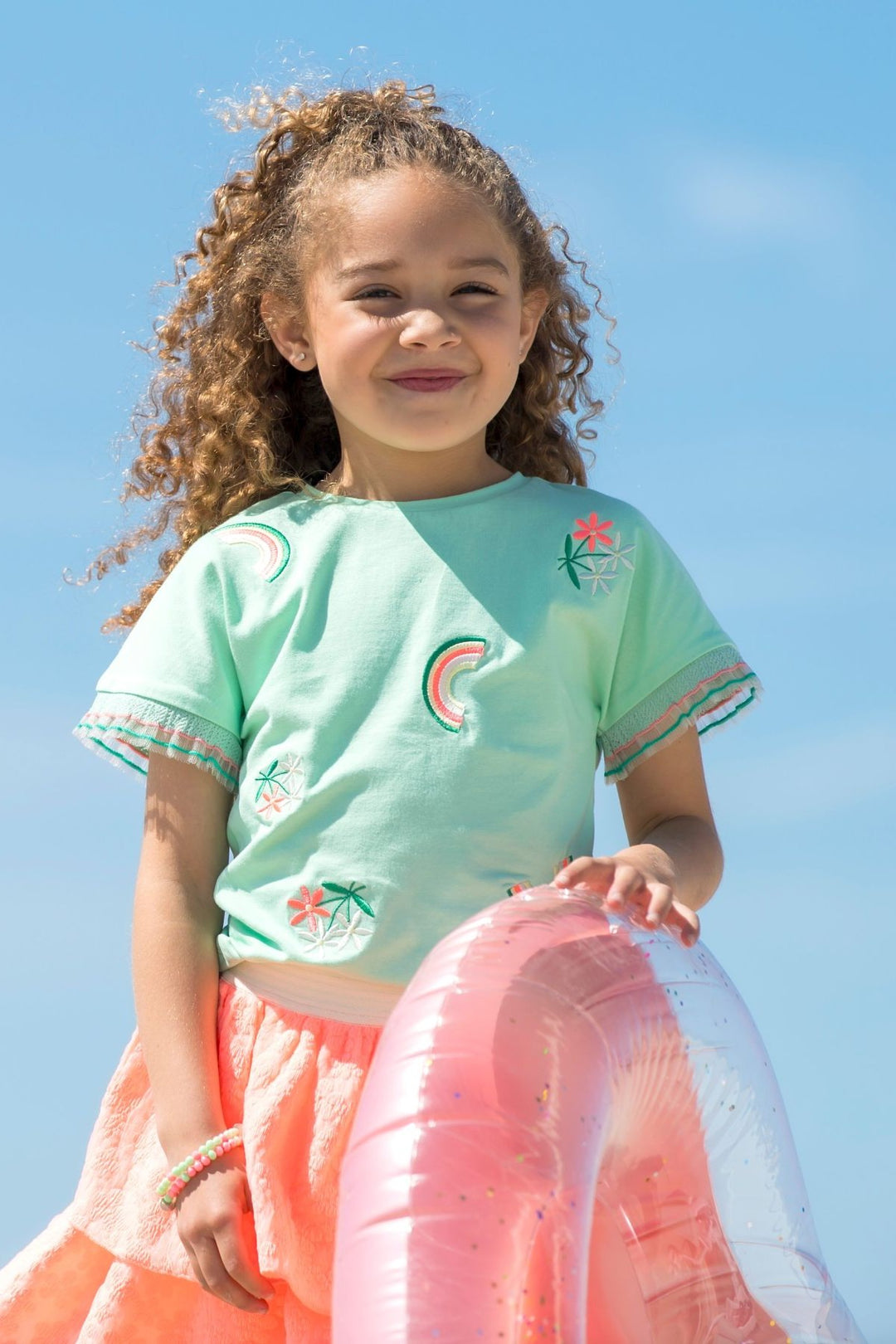 A young girl wearing a mint green short-sleeve t-shirt with colorful embroidered motifs and a peach skirt, holding a pink inflatable pool float against a clear blue sky.