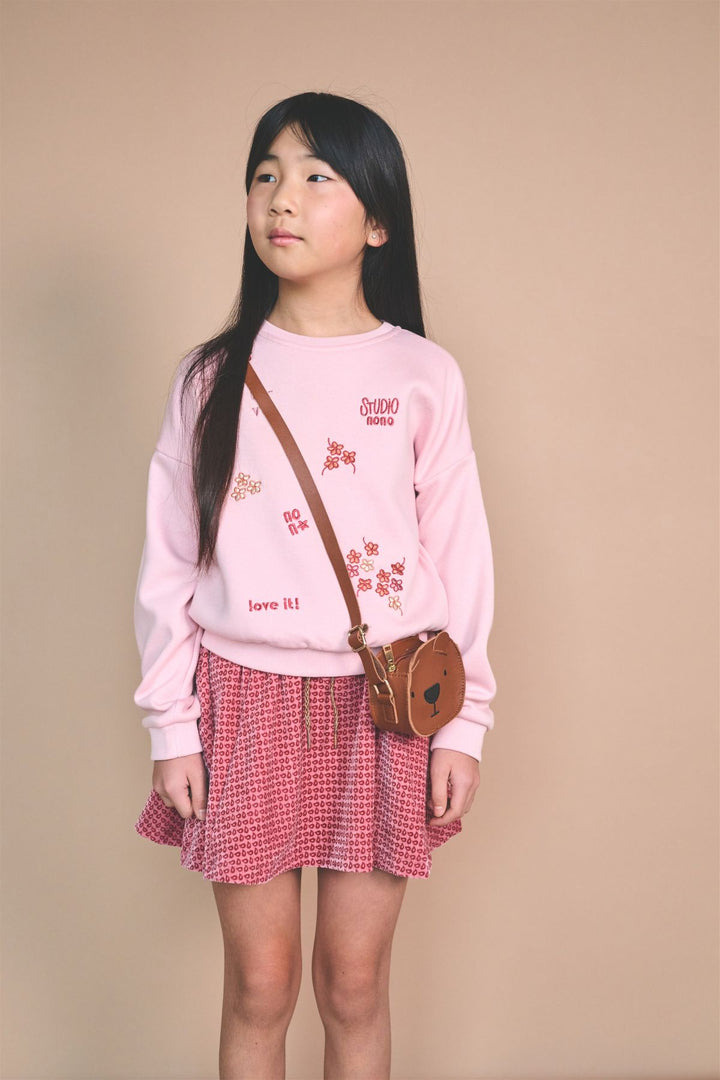 A young girl with long black hair stands against a solid beige background, wearing a light pink sweatshirt with cherry blossom graphics, a red and white gingham pleated skirt, and carrying a brown leather crossbody bag.