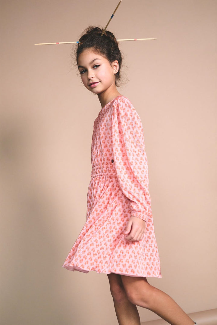 A young girl with her hair in a high bun wearing a pink and white patterned long-sleeve dress, posing against a solid beige studio background.