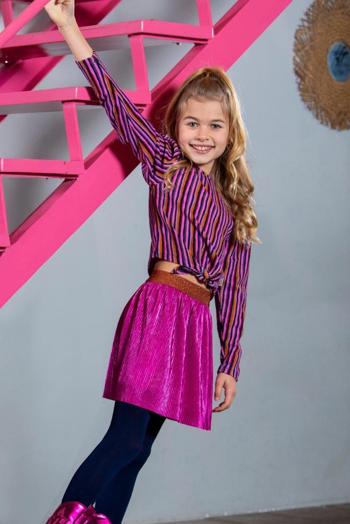 Young girl in a striped shirt and pink skirt standing against a gray wall with a pink staircase.