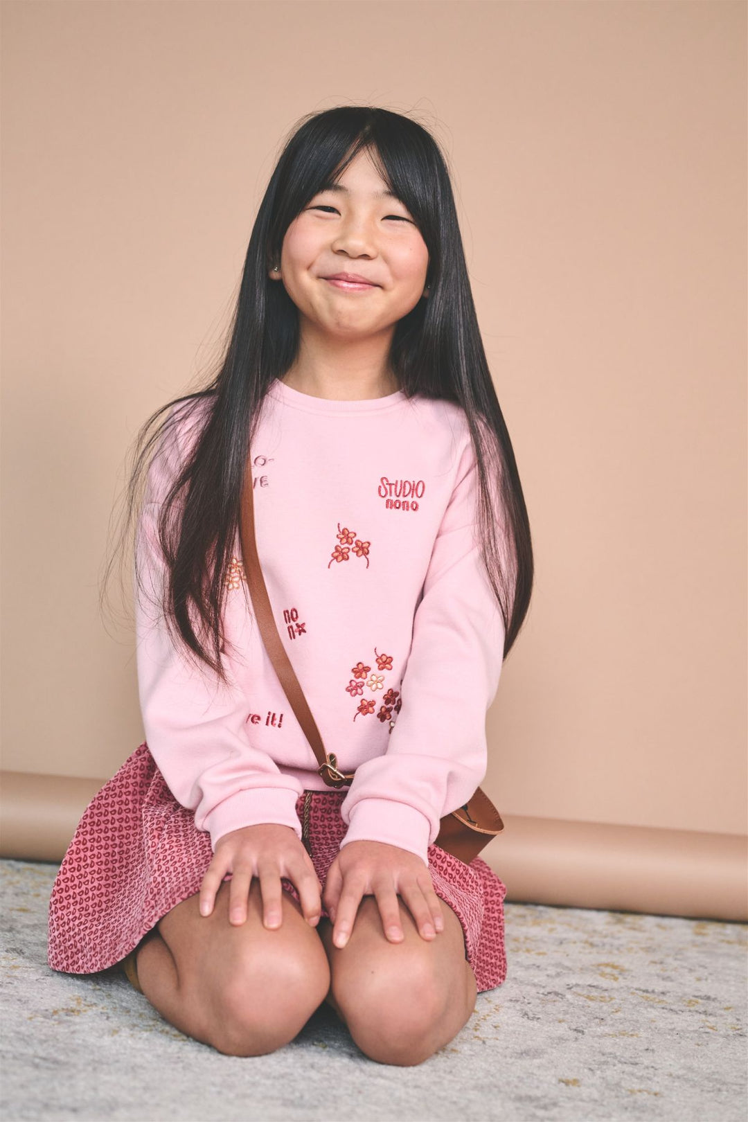 A young girl with long black hair sits cross-legged on a textured gray floor against a solid beige backdrop. She is wearing a light pink crewneck sweatshirt with red cherry blossom graphics and text, paired with a red and white checkered skirt and a brown crossbody bag.