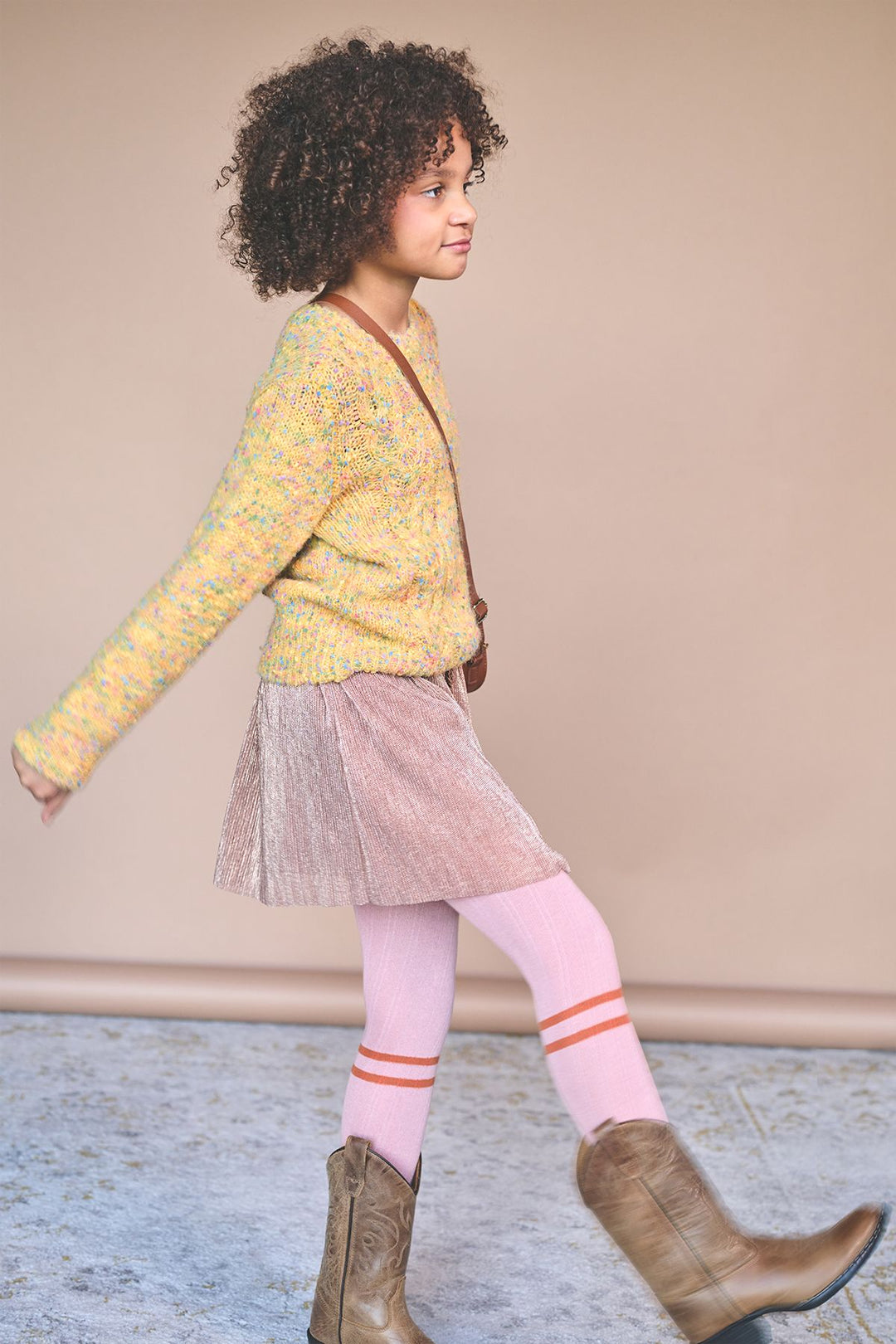 A young girl with curly dark hair is shown in profile wearing a yellow knit cardigan, a pink pleated skirt, light pink striped tights, and brown cowboy boots against a solid beige background.
