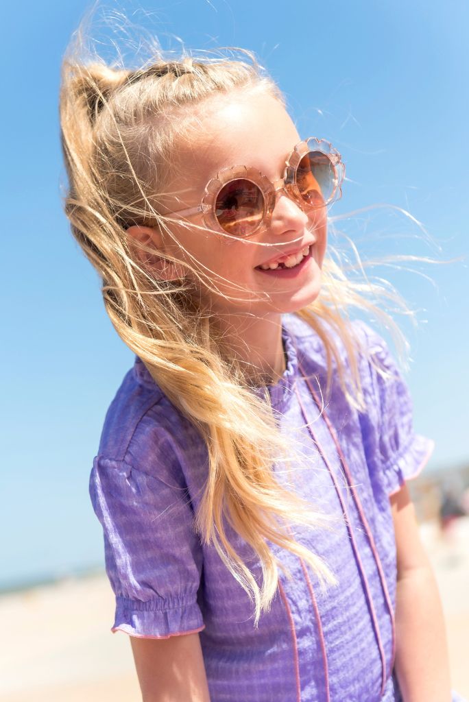 Young girl with blonde hair wearing sunglasses and the purple dress Dotty against a clear blue sky.