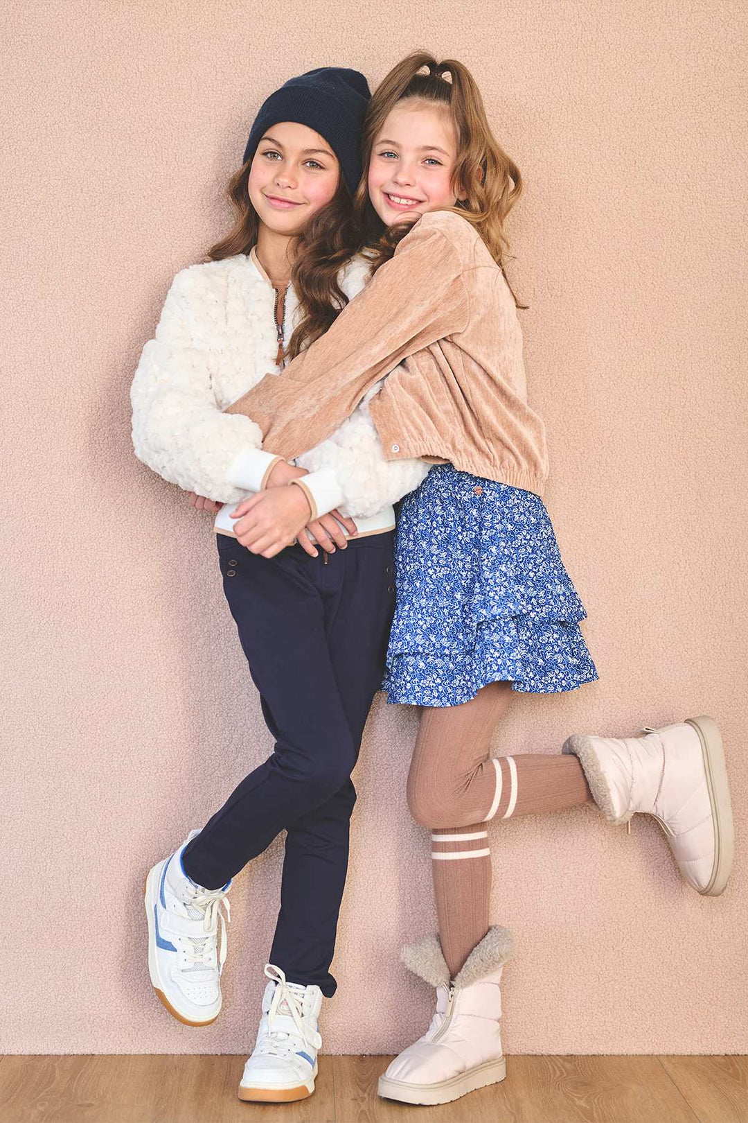 Two young girls hugging and posing together in a studio setting against a solid beige-pink wall. The girl on the left wears a black beanie, a white fuzzy jacket, dark navy pants, and white sneakers.