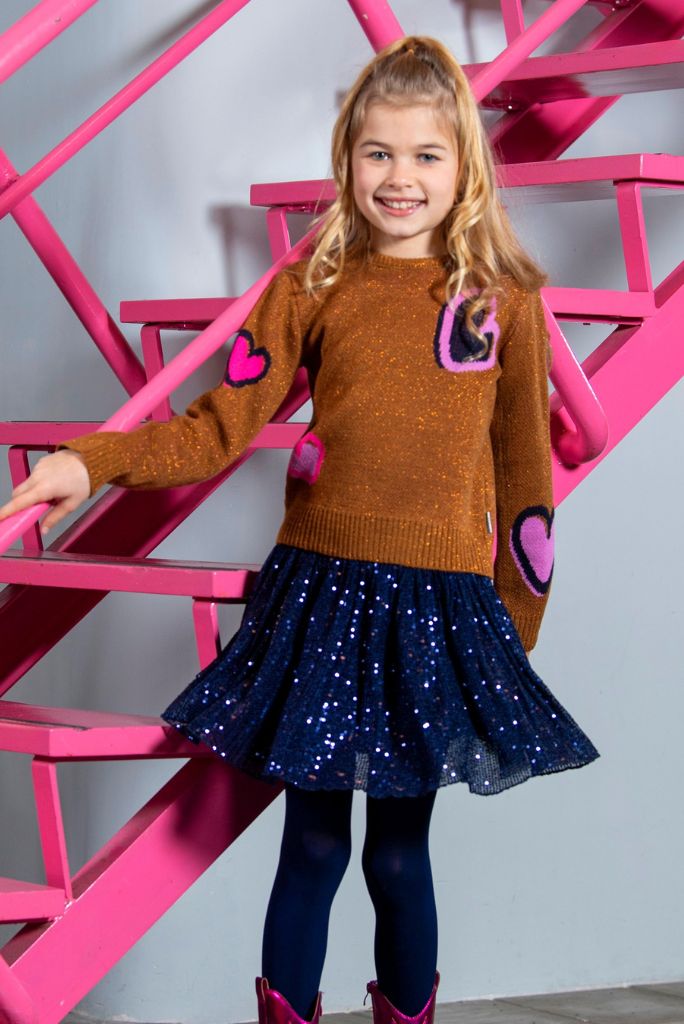 Young girl wearing a brown sweater with heart designs and a navy blue sequin skirt standing against a pink metal structure.