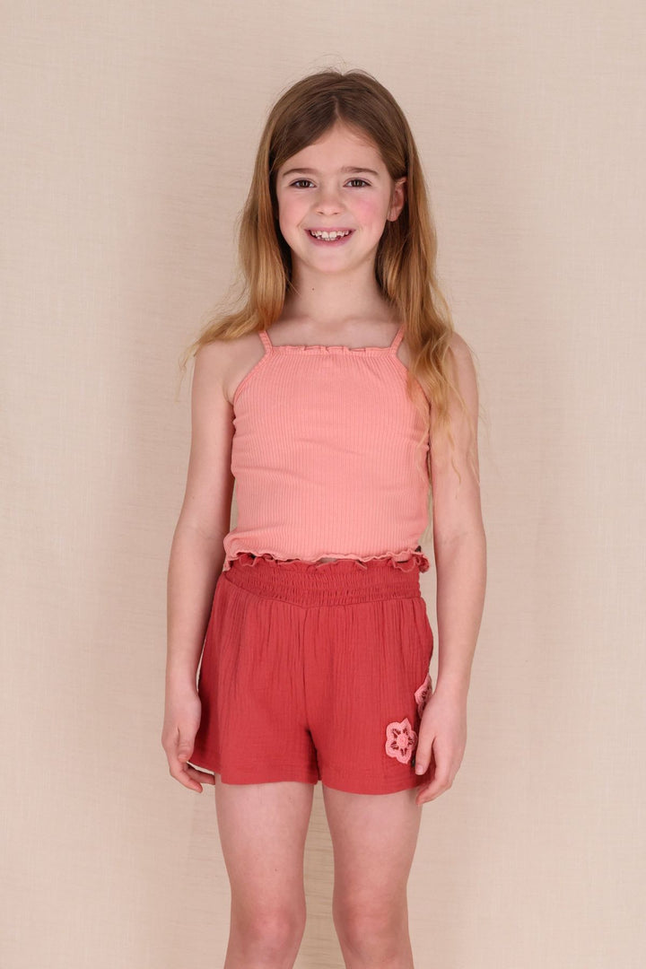 A young girl wearing a coral pink ribbed singlet and matching coral red shorts with a small embroidered flower on the left leg.