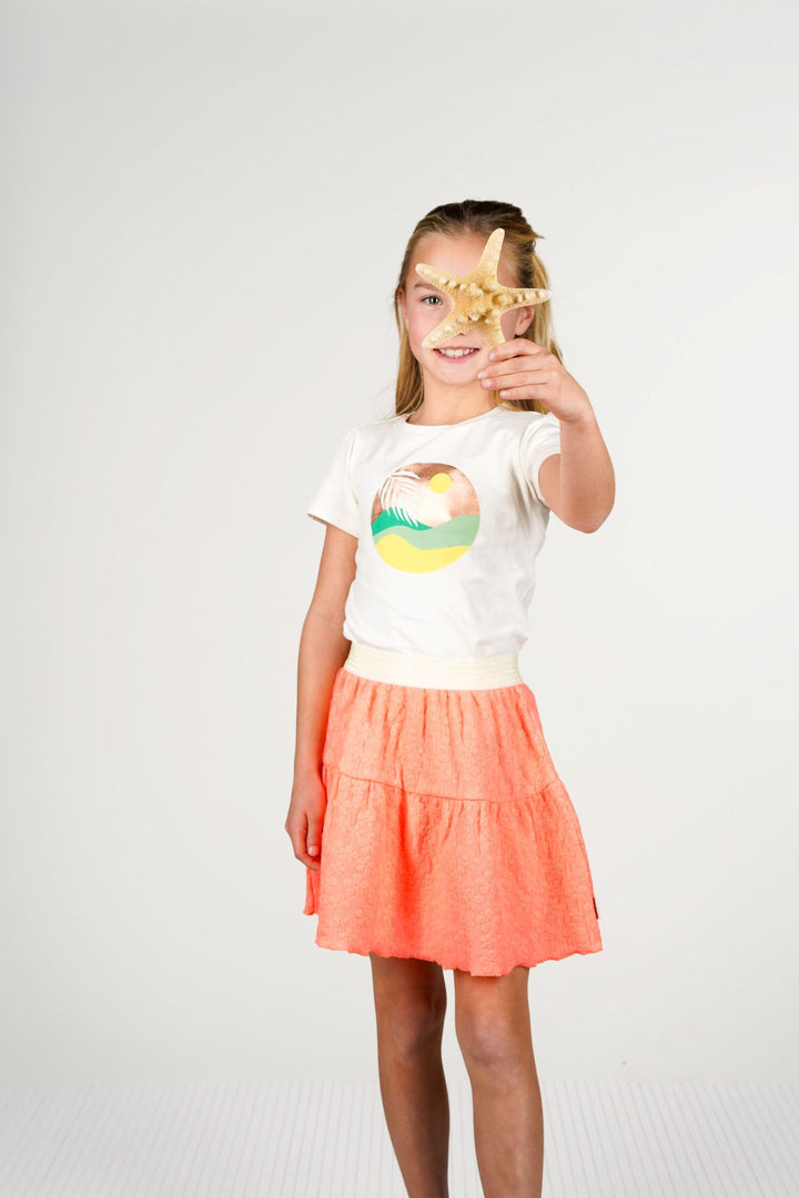A young girl stands against a light gray studio background wearing a white graphic t-shirt and a tiered coral-orange skirt. She holds a starfish in front of her eyes.