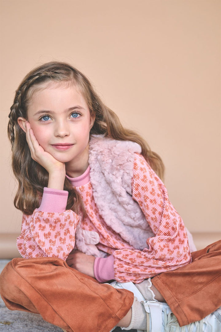 A young girl with long light brown hair sits cross-legged against a solid beige background, wearing a pink patterned long-sleeve top, a fuzzy light pink vest, rust-brown pants, and white sneakers.