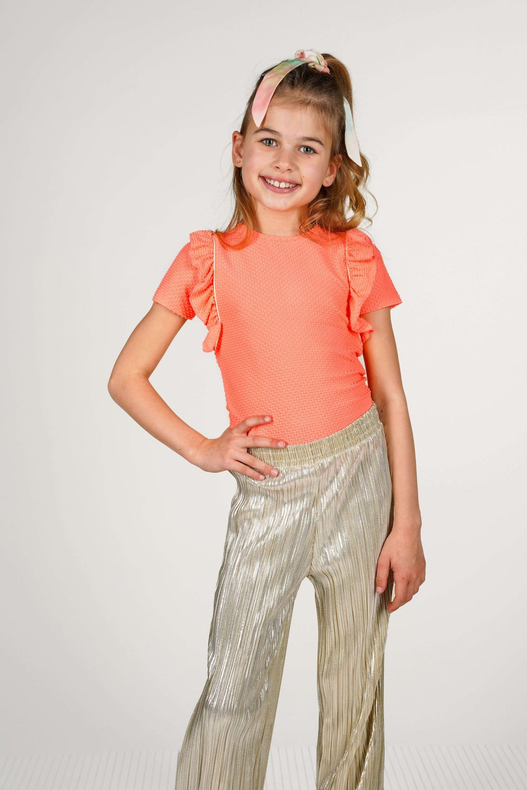 A young girl in a short-sleeve coral orange top with ruffle details and shiny metallic gold pleated pants, standing against a light gray studio background.
