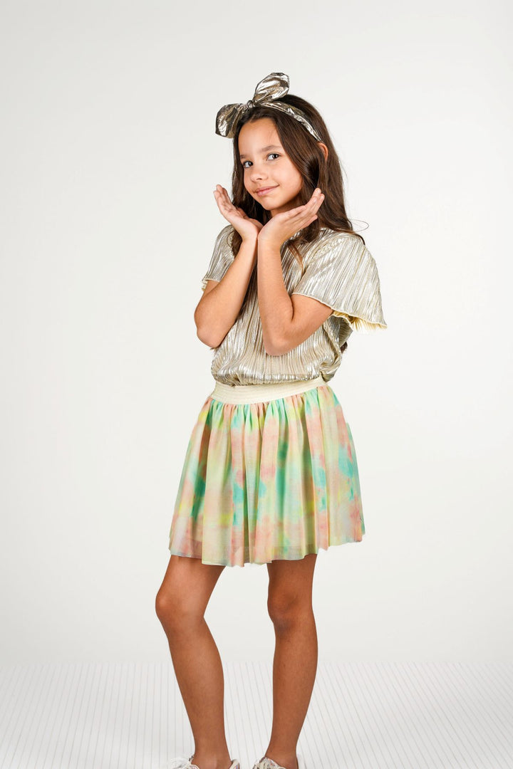 A young girl modeling a two-piece outfit consisting of a metallic gold pleated short-sleeve top and a pastel tie-dye pleated mini skirt in green, yellow, and pink, styled with a matching metallic bow headband against a white studio background.