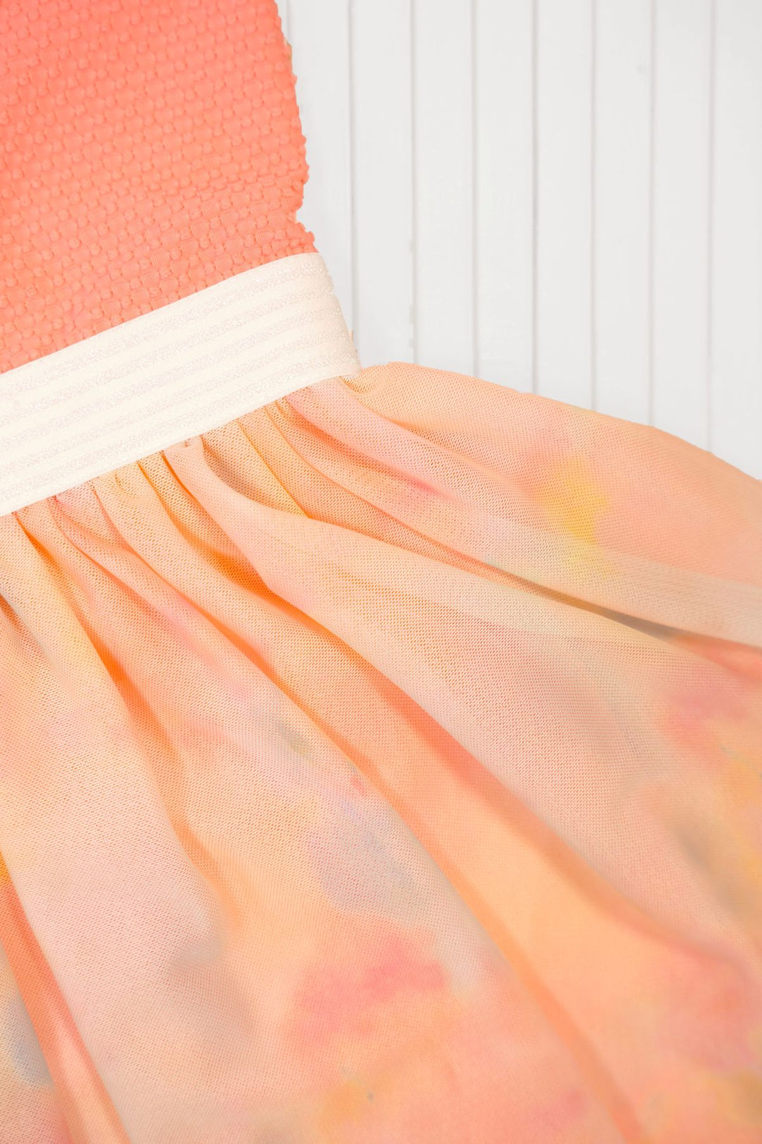 A close-up of a peach and orange tulle skirt with a textured coral-pink bodice and a white elastic waistband, set against a white paneled background.
