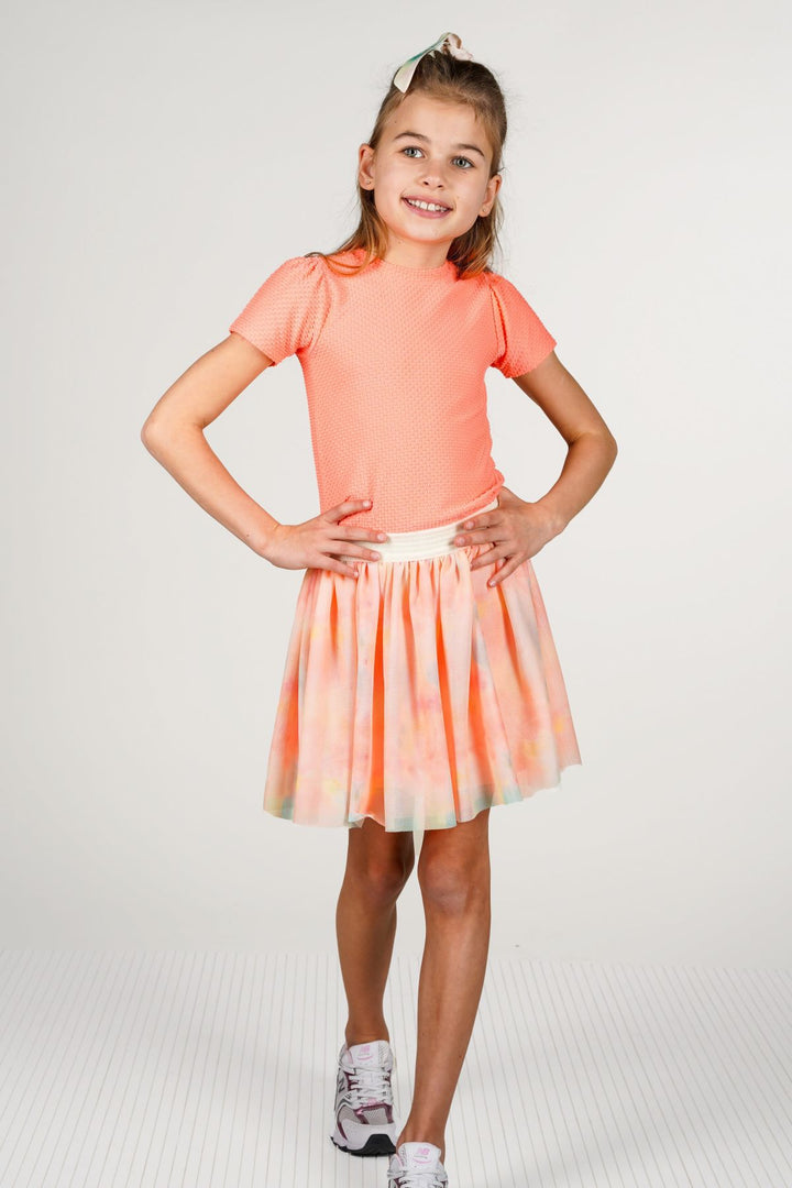 A young girl modeling a dress with a solid coral short-sleeve top and a pleated tulle skirt with a peach, orange, and pale green tie-dye pattern, standing against a light gray studio background.