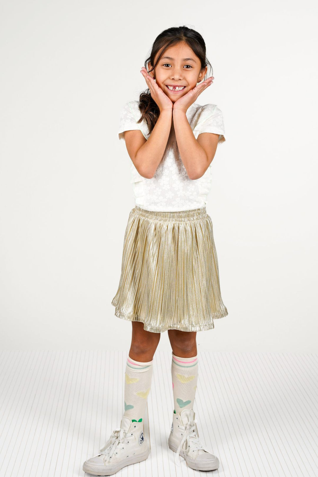 A young girl posing in a white short-sleeve top and a shiny gold pleated skirt, paired with white knee-high socks and white sneakers, against a plain white studio background.