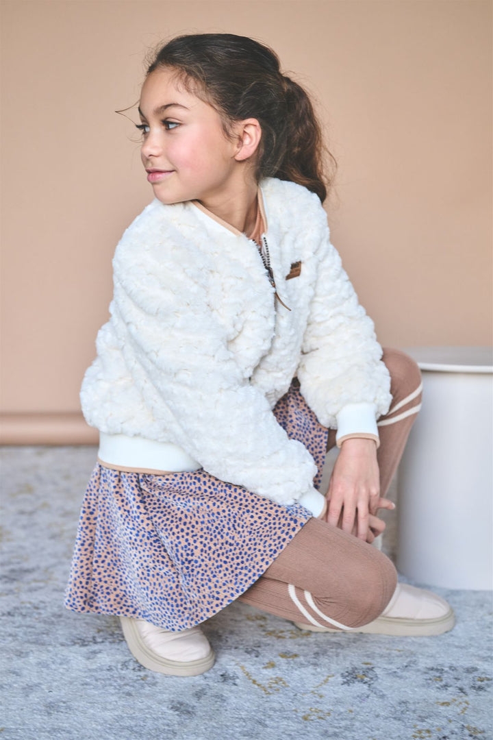 A young girl with her hair in a ponytail is crouching in a studio setting. She is wearing a white fuzzy cropped jacket, a patterned skirt with blue and orange dots, beige tights, and cream-colored shoes.