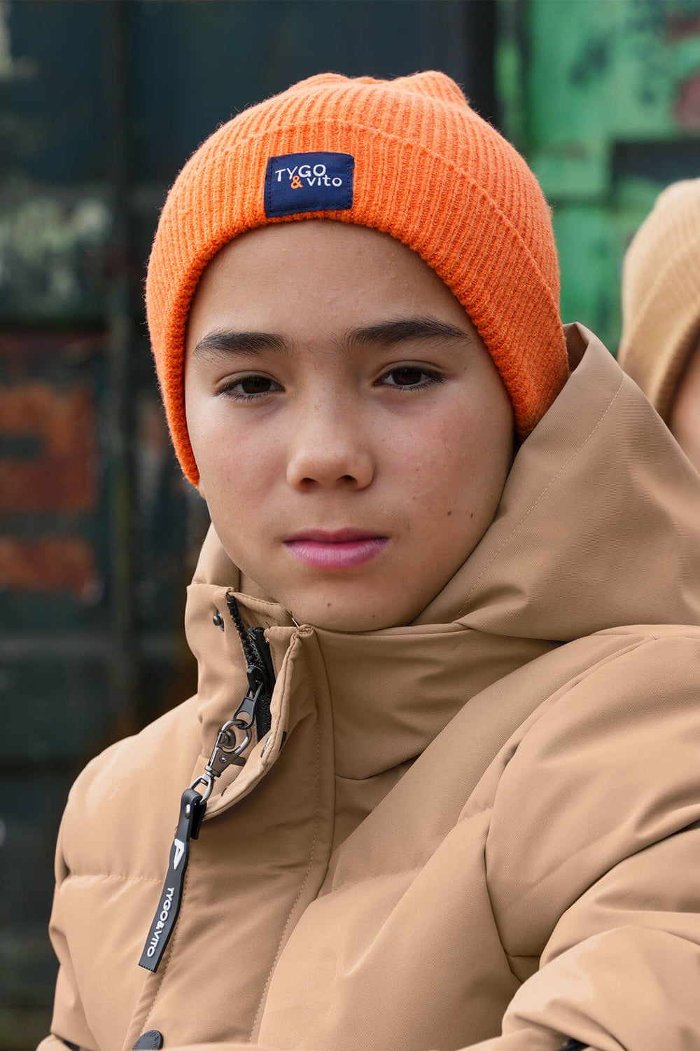 A boy wearing an orange beanie with a visible brand logo, standing outdoors.